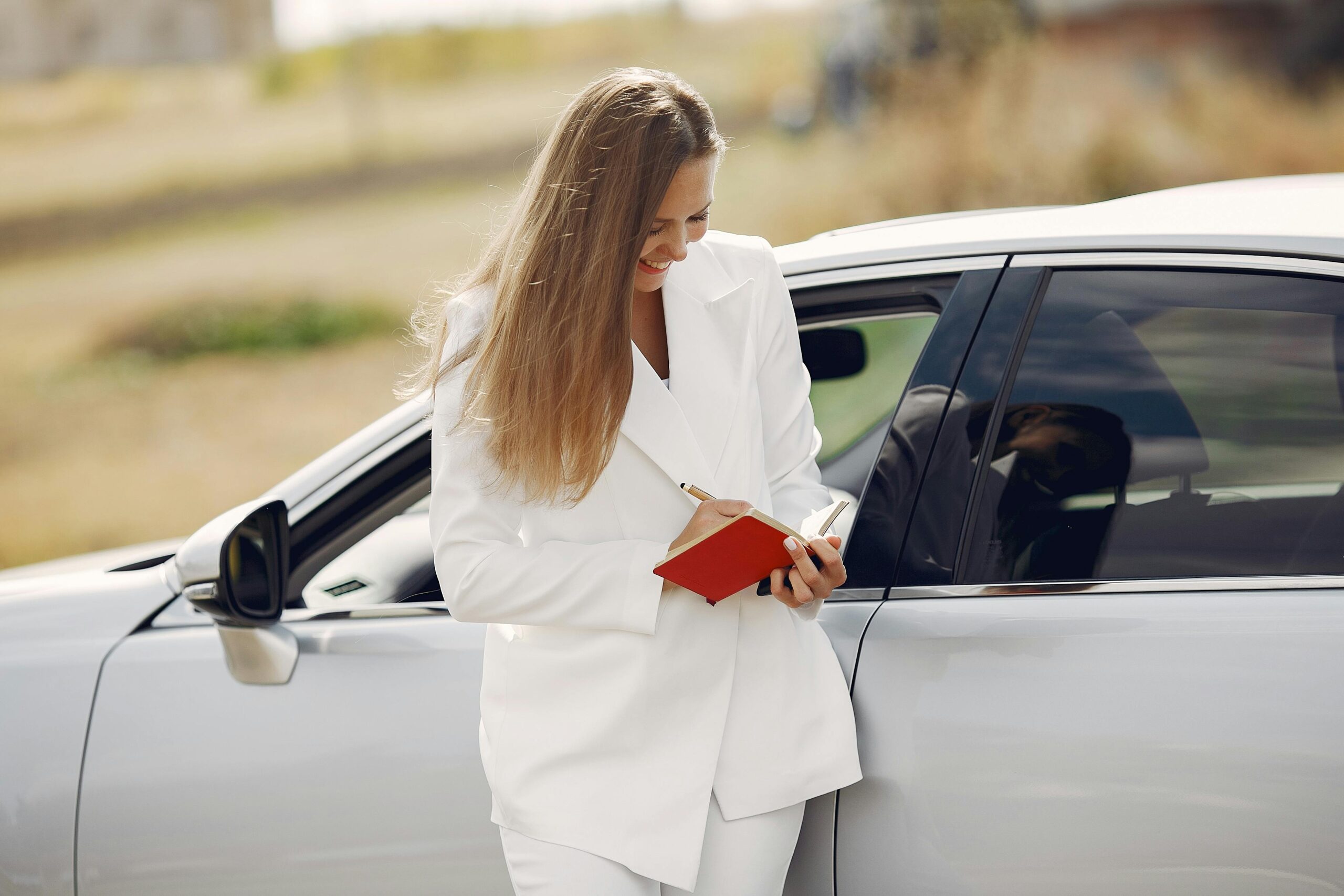 Elegant young female traveler wearing white suit leaning on car while writing information in notebook during business trip in sunny summer day in countryside